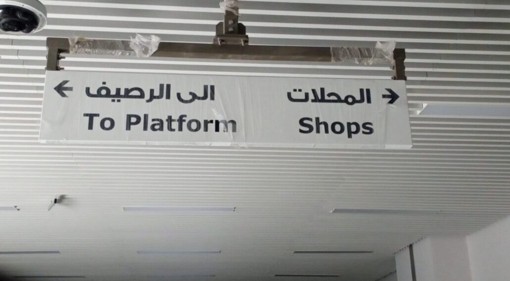 Directional sign in a metro station with Arabic and English text, guiding passengers to the platform and shops. Clear wayfinding signage for public transit.